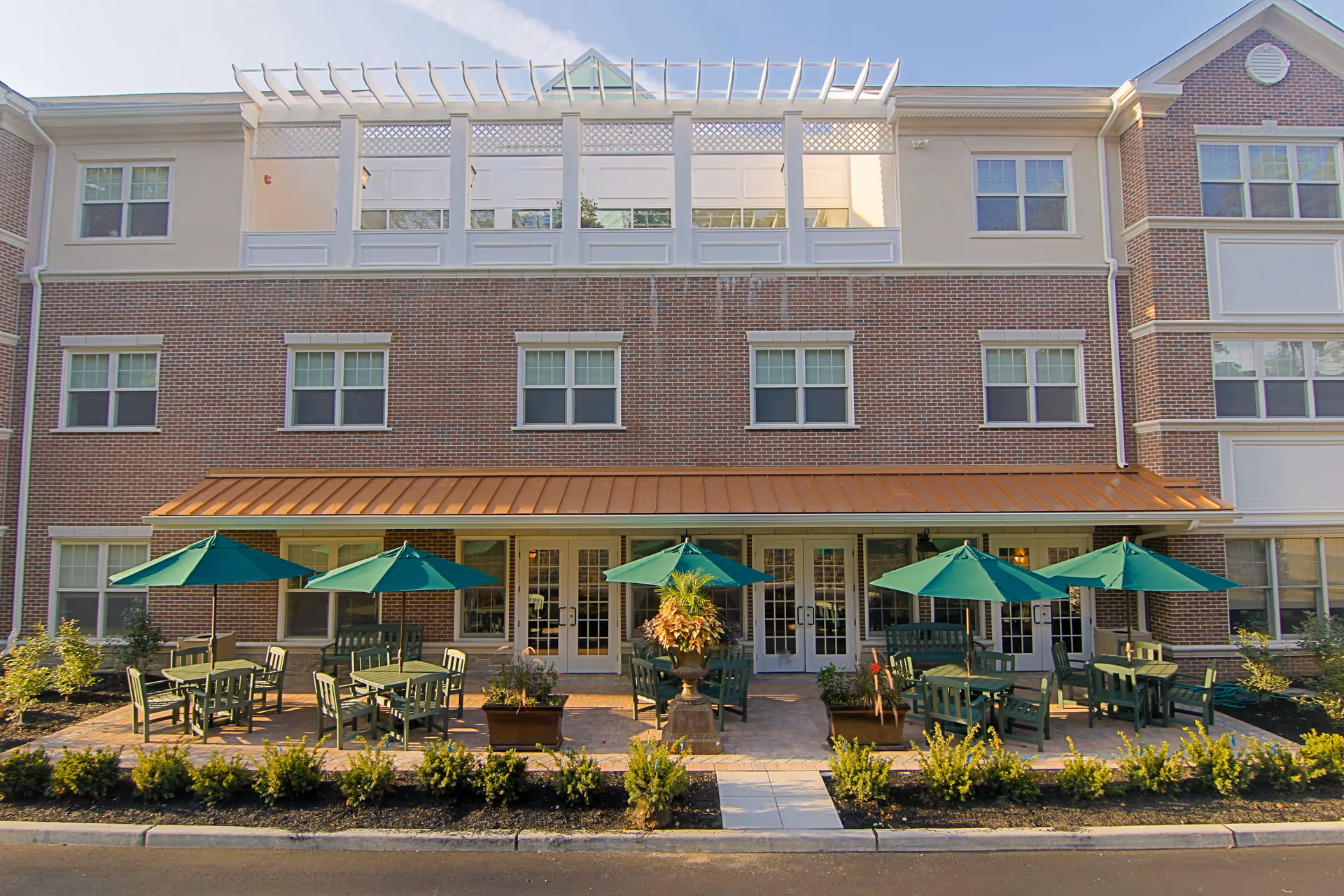 Outdoor patio area of a senior living facility with several green tables and chairs under green umbrellas, in front of a three-story brick building with multiple windows and a copper-colored awning.