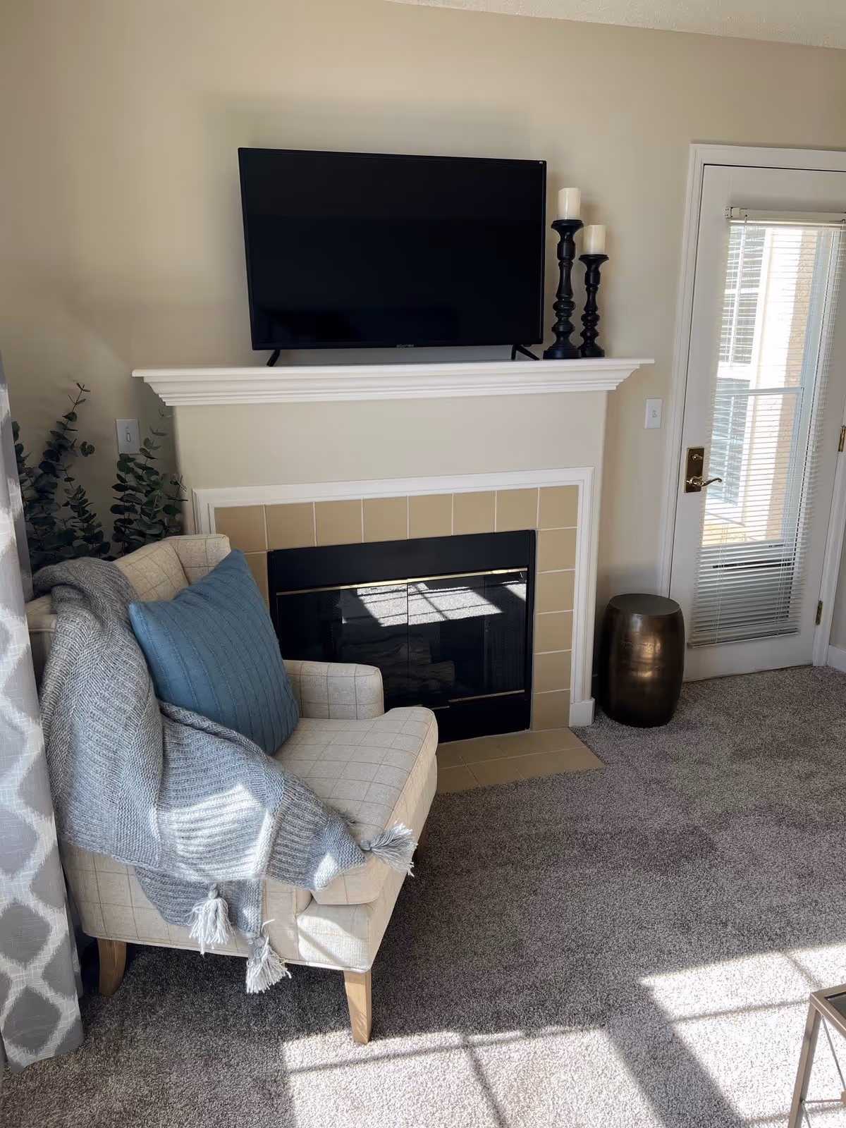 A cozy living room corner featuring a beige armchair with a blue pillow and a gray knitted throw draped over it. Behind the chair is a fireplace with a beige tiled surround and a white mantel. On the mantel sits a flat-screen TV and three black candle holders with white candles. To the right is a glass door with white blinds, and a small metallic side table is positioned next to the door. The floor is carpeted in a light gray color.