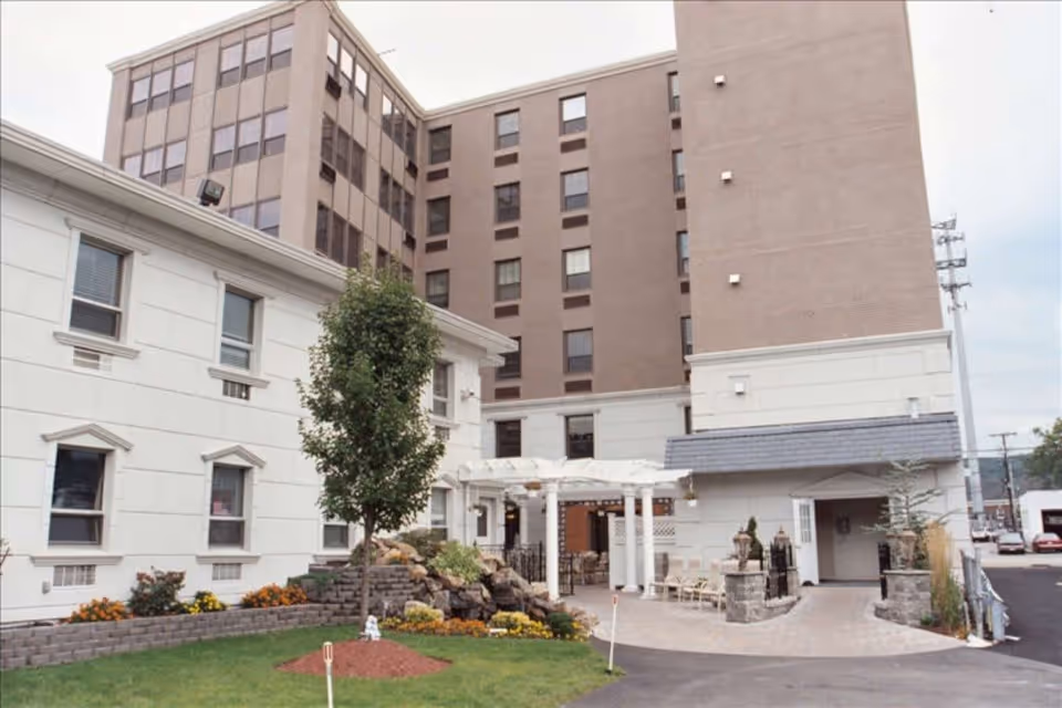Exterior view of Excelcare at Dover facility showing a multi-story building with a white and beige facade, a small landscaped garden with a tree and flowers, and a covered outdoor seating area with chairs and tables.