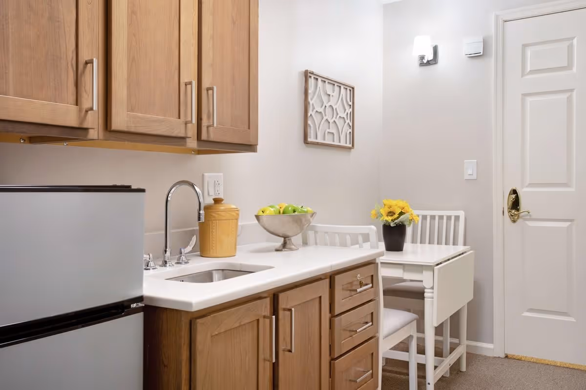 A compact kitchenette with wooden cabinets, a sink and small refrigerator next to a white dining nook with a vase of flowers.