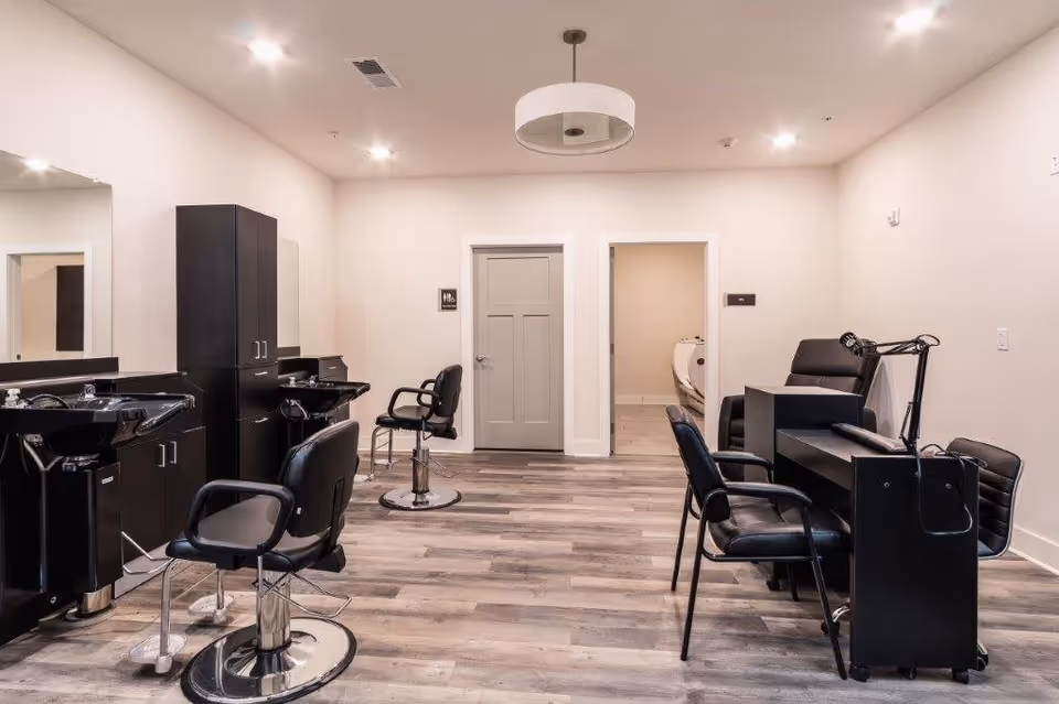 Interior view of a salon area in a senior living facility with black salon chairs, sinks for hair washing, a manicure station with black chairs, light wood flooring, white walls, and a modern ceiling light fixture. Two doors are visible in the back wall, one labeled as a restroom.