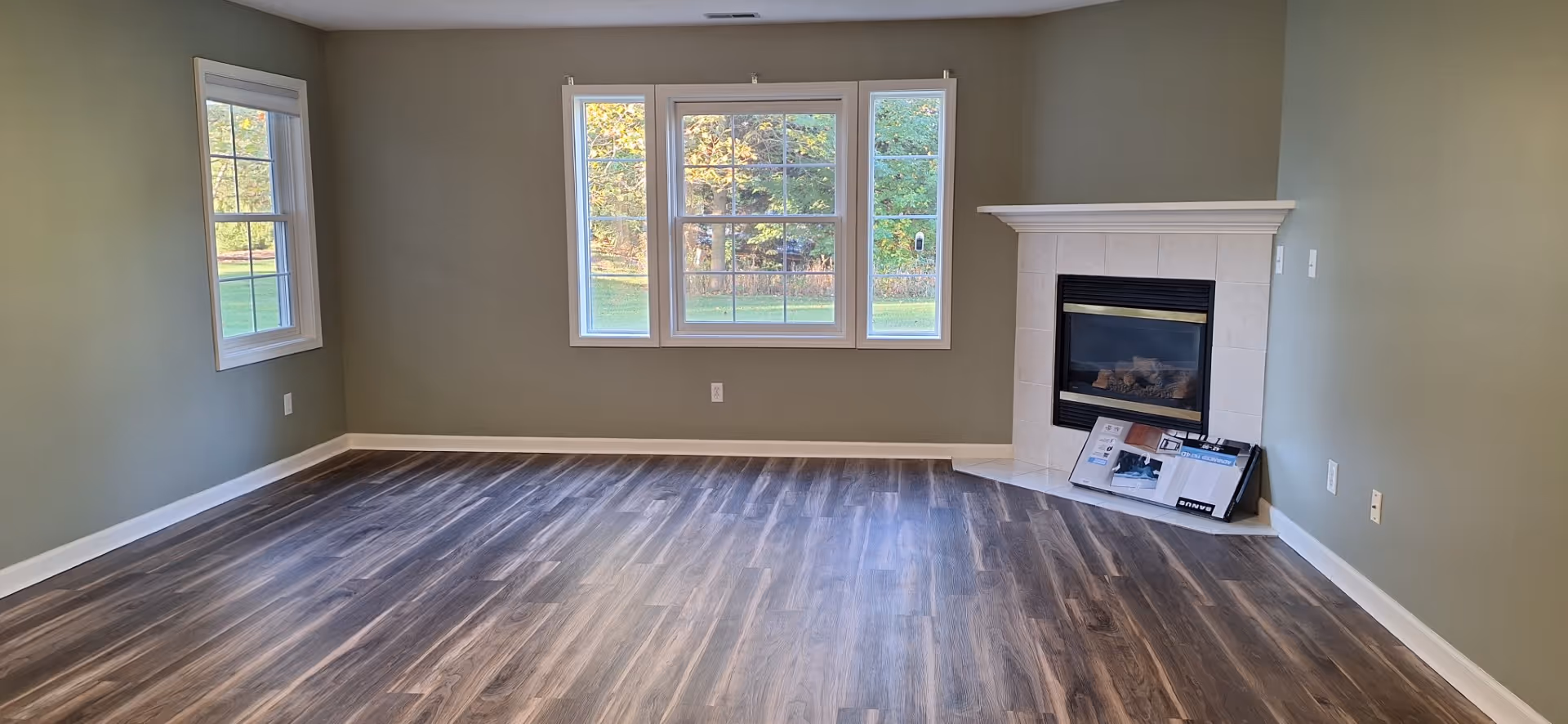 Empty room with green walls, three large windows showing an outdoor view with trees, wood-patterned flooring, and a corner fireplace with a tiled surround. There are some boxes placed on the hearth of the fireplace.