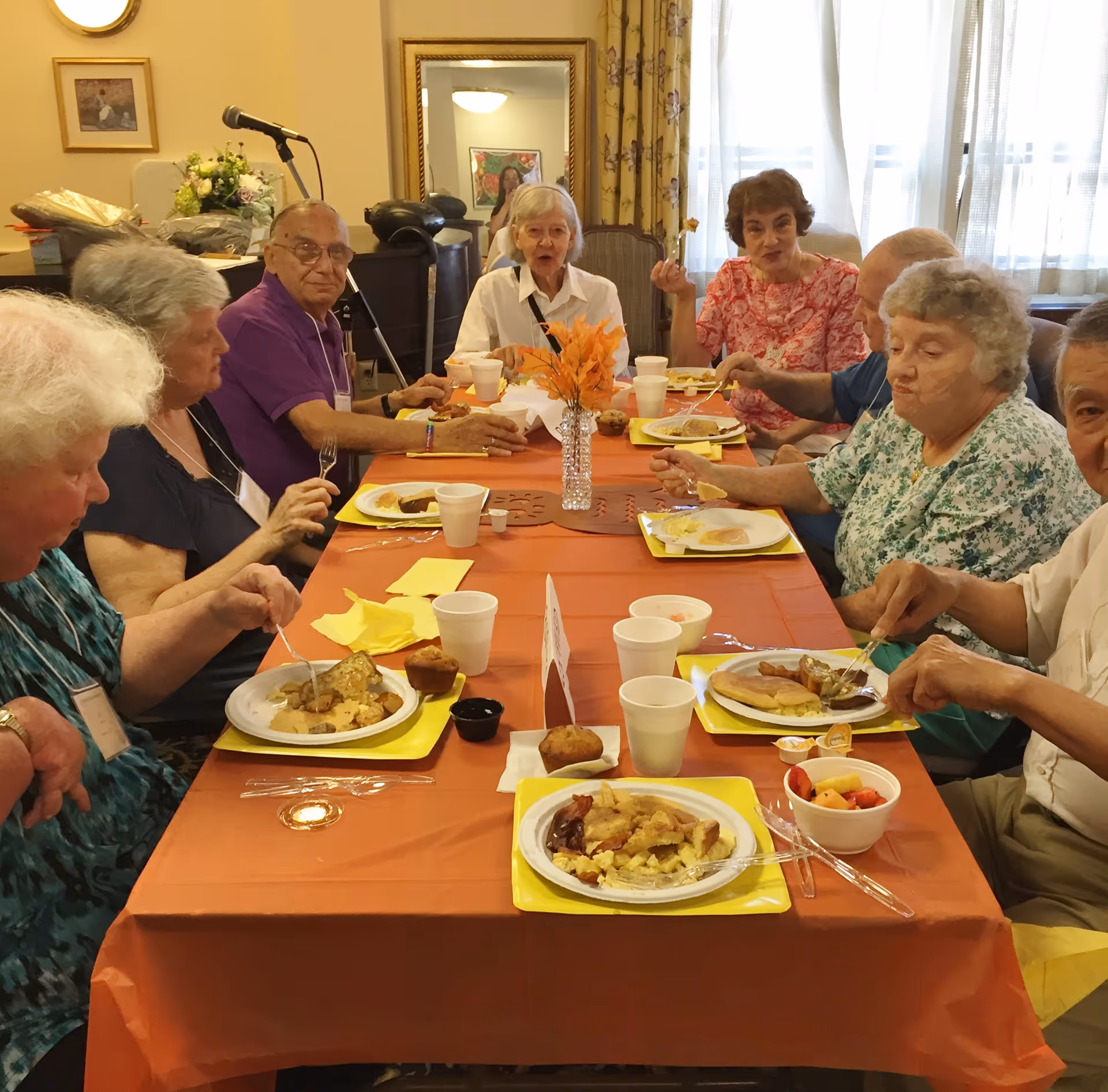 A group of elderly people sitting around a long table covered with an orange tablecloth, eating a meal together in a dining room. The table is set with yellow trays, white disposable cups, plates of food, and a small vase with orange flowers in the center. The room has a microphone stand, framed pictures on the wall, and a large window with sheer curtains.