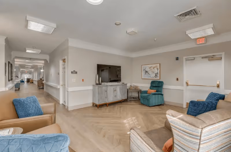 A bright and spacious common area in a senior living facility with comfortable armchairs in blue, tan, and striped upholstery arranged around a TV on a wooden cabinet. The room has light-colored walls, a framed floral artwork, and a long hallway extending into the distance with additional seating and artwork on the walls.