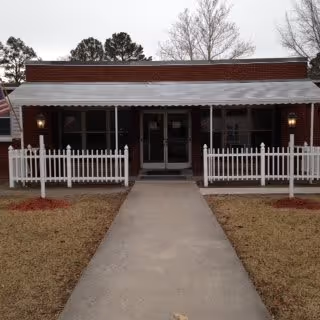 Front entrance of a single-story brick building with a covered porch, white picket fence, and a concrete walkway.