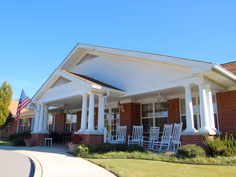 Front exterior view of a single-story brick building with a white covered porch supported by white columns. Several white rocking chairs are arranged on the porch. An American flag is displayed near the entrance. The sky is clear and blue.