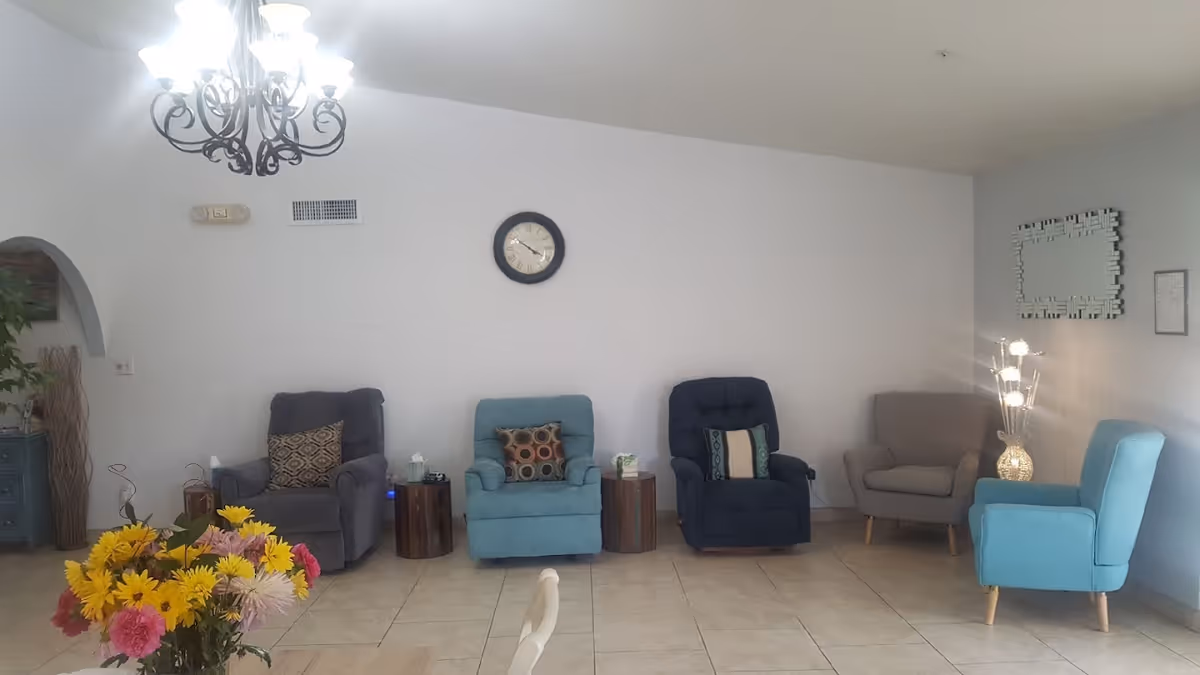 A cozy living room area with five armchairs in different colors arranged against a white wall. There are two small wooden side tables with tissue boxes between the chairs. A decorative wall clock is centered above the chairs. A chandelier with multiple lights hangs from the ceiling. A vase with colorful flowers is visible in the foreground on a table. A decorative floor lamp and a mirror are on the right side wall.