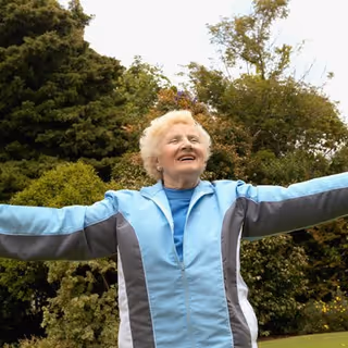 An elderly woman wearing a light blue and gray jacket stands outdoors with her arms outstretched, smiling joyfully in front of green trees and bushes.