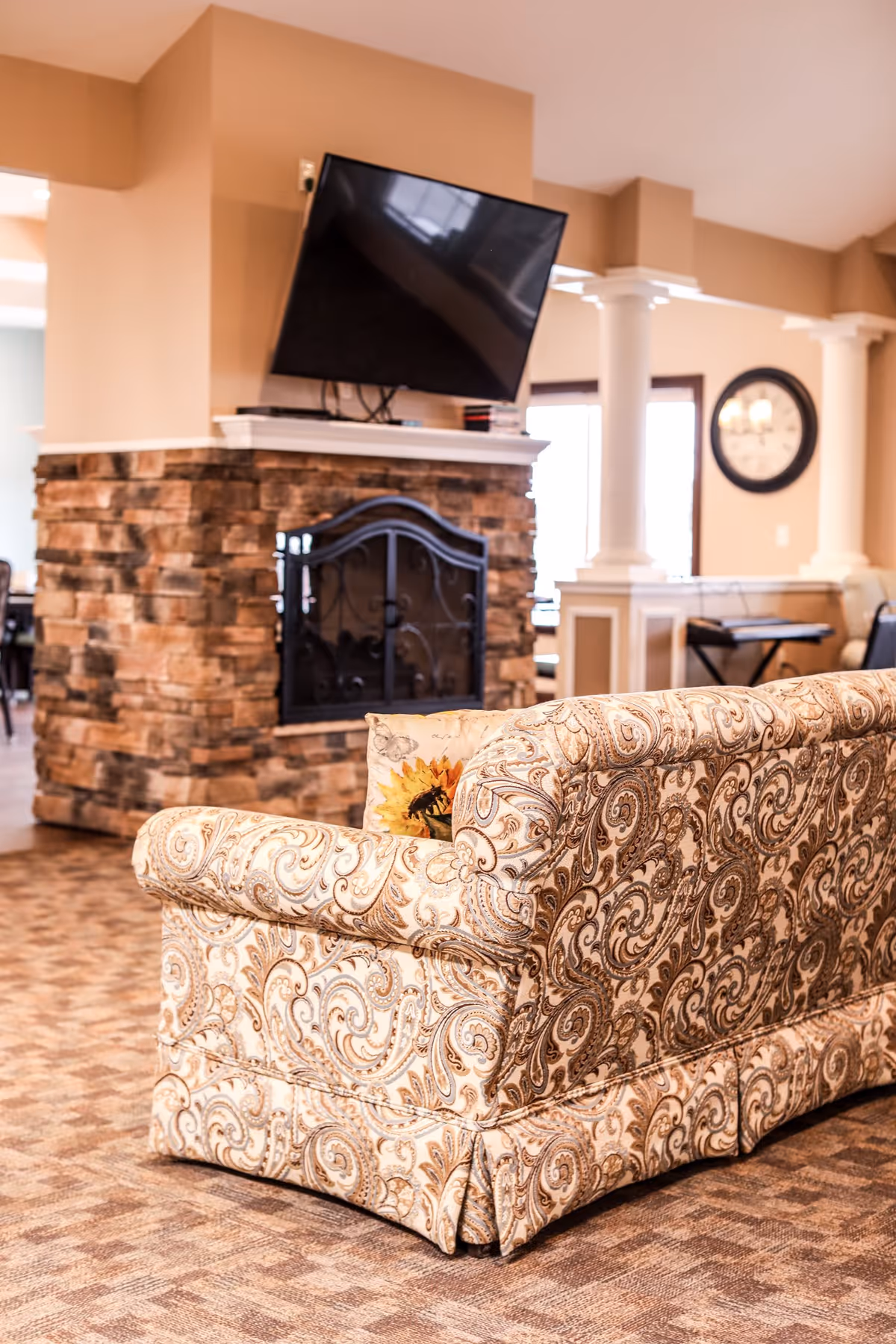 A cozy living room area featuring a patterned beige and brown sofa with a sunflower decorative pillow. In the background, there is a stone fireplace with a black metal screen and a flat-screen TV mounted above it. The room has beige walls, white columns, and a large clock on the wall near a window letting in natural light.