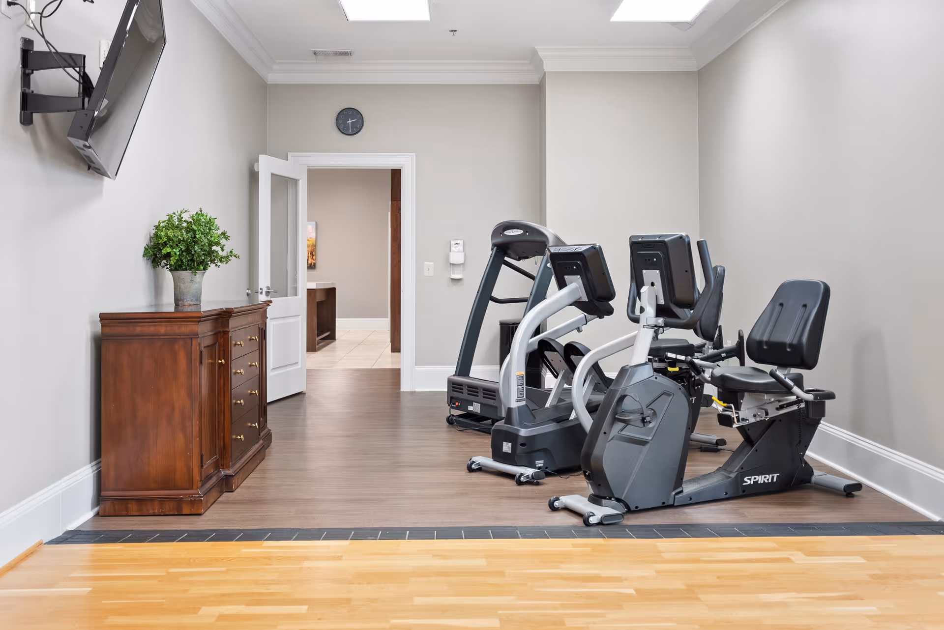 A small fitness room with exercise equipment including a recumbent bike and a treadmill. The room has light gray walls, wood flooring, a wooden cabinet with a potted plant on top, a wall-mounted TV, and an open door leading to another area with tiled flooring.