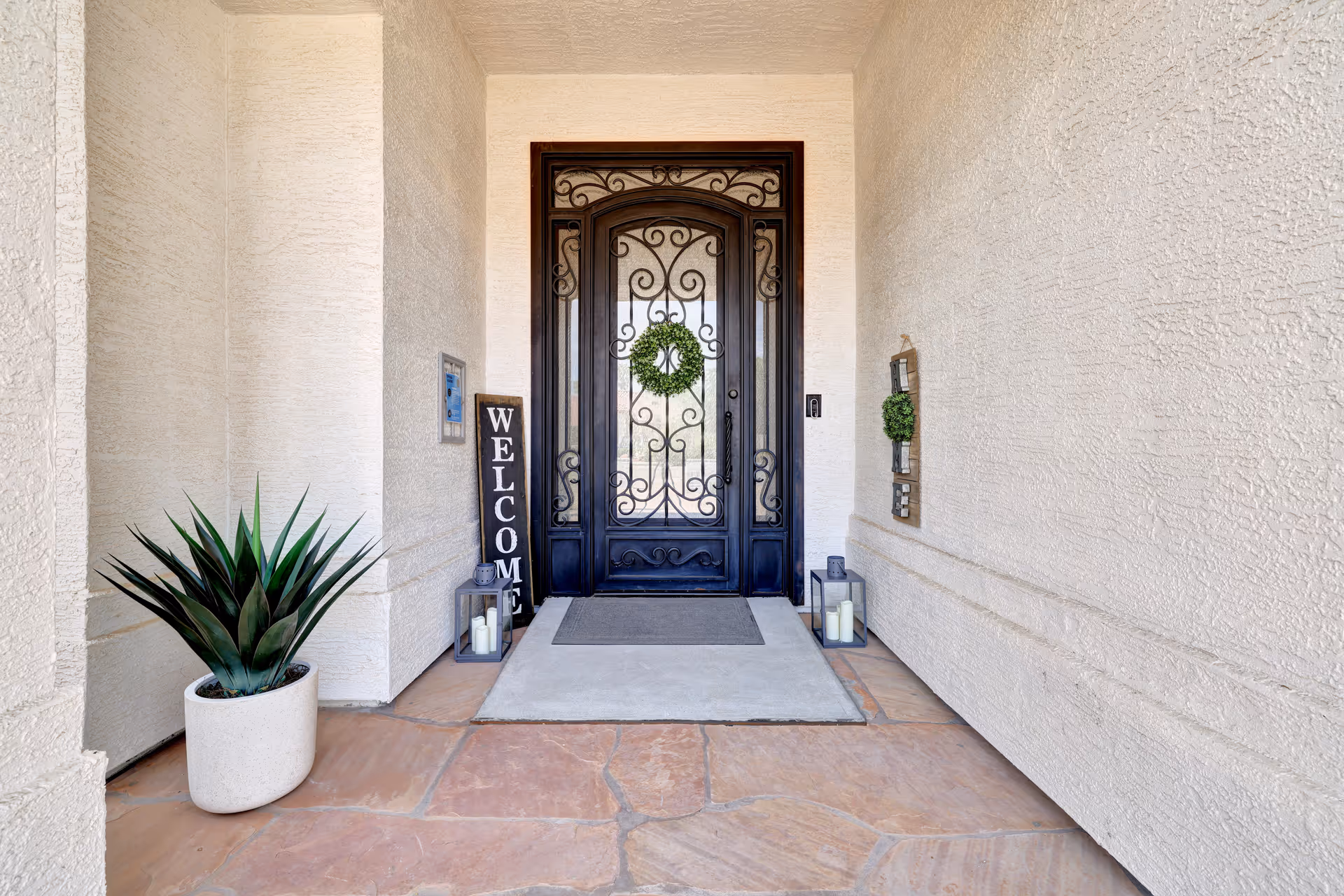 Covered front entry with a decorative wrought-iron door, a 'WELCOME' sign, potted plant, wreath, and lantern candles on the porch.