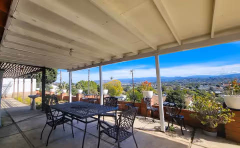 Covered outdoor patio area with a metal table and several chairs. The patio overlooks a scenic view of a city with trees and mountains in the distance. There are potted plants along the edge of the patio.