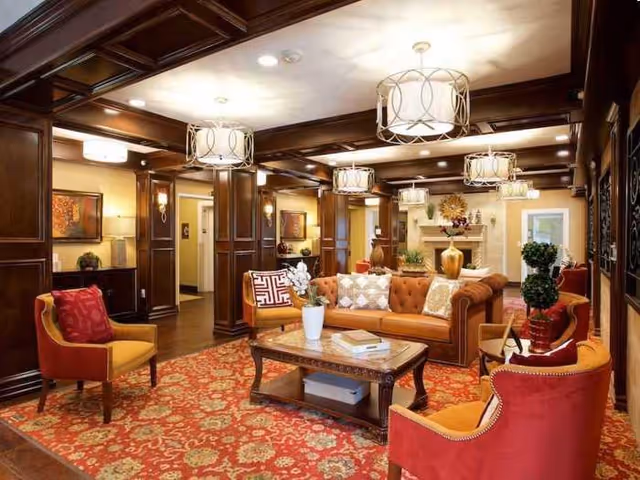 Elegant communal living room with a tufted leather sofa and armchairs around a coffee table on a patterned rug, wood-paneled columns, coffered ceiling, and multiple pendant lights.