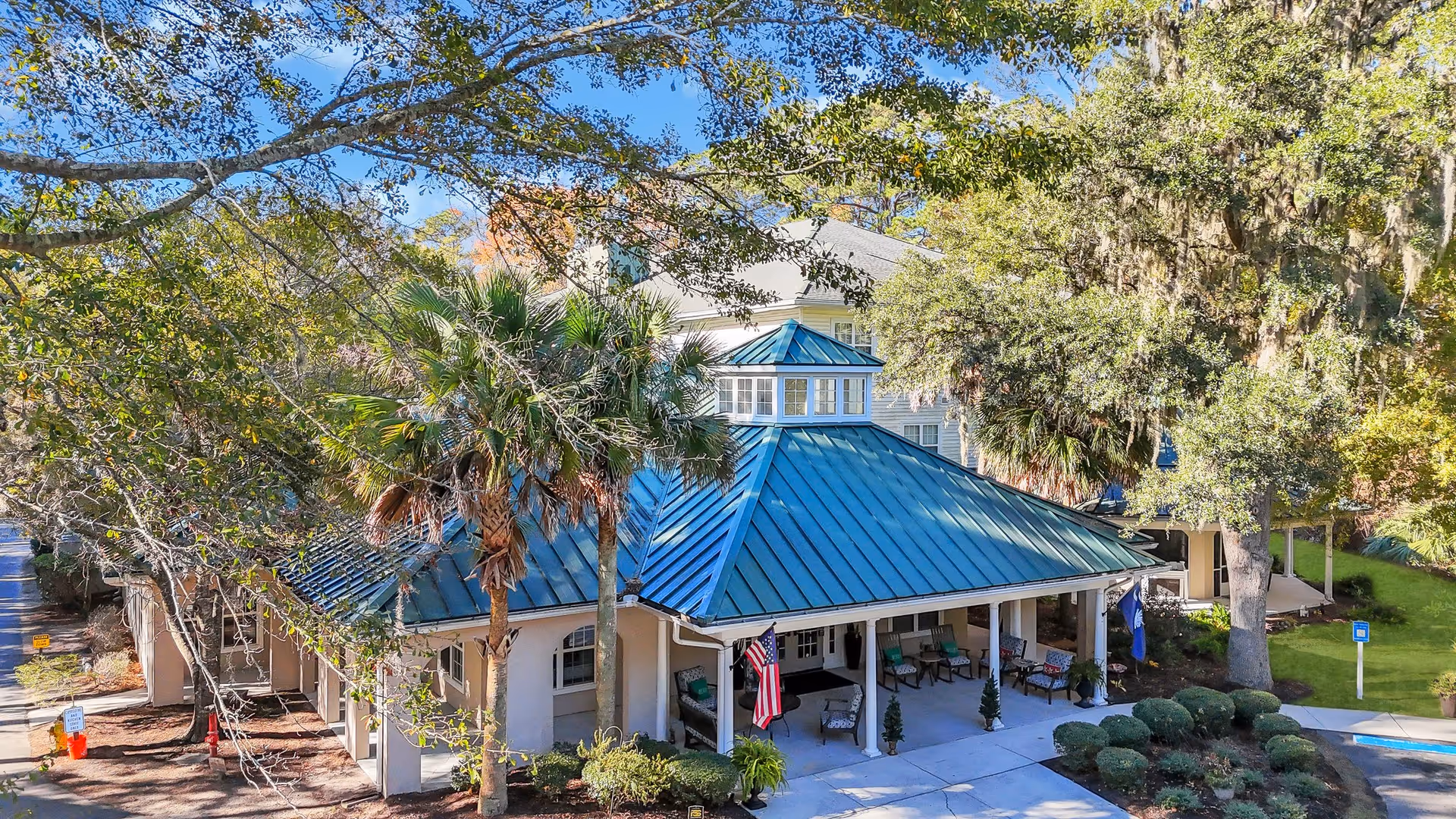 Front exterior of a single-story building with a blue metal roof, covered porch and surrounding trees and landscaping.