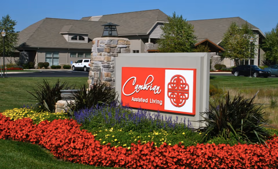 Exterior view of Cambrian Assisted Living facility with a large sign in front surrounded by colorful flower beds and green grass. The building is beige with a sloped roof, and there are a few cars parked in the background.
