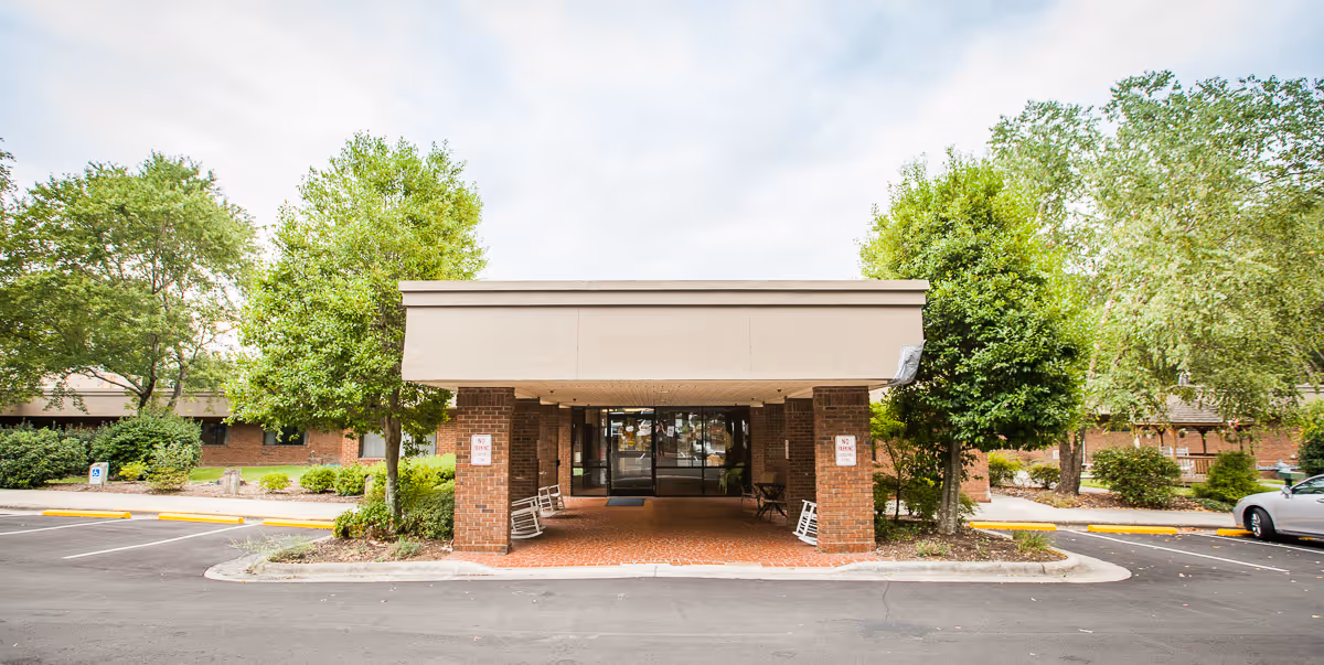 Front entrance of a single-story brick building with a covered drop-off area supported by brick pillars. There are rocking chairs placed under the covered area, surrounded by green trees and shrubs. The parking lot is visible in the foreground with a few parked cars.