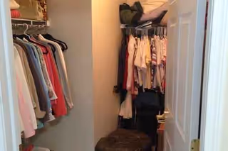 Narrow closet filled with hanging clothes, shelves, and a stool near the doorway.