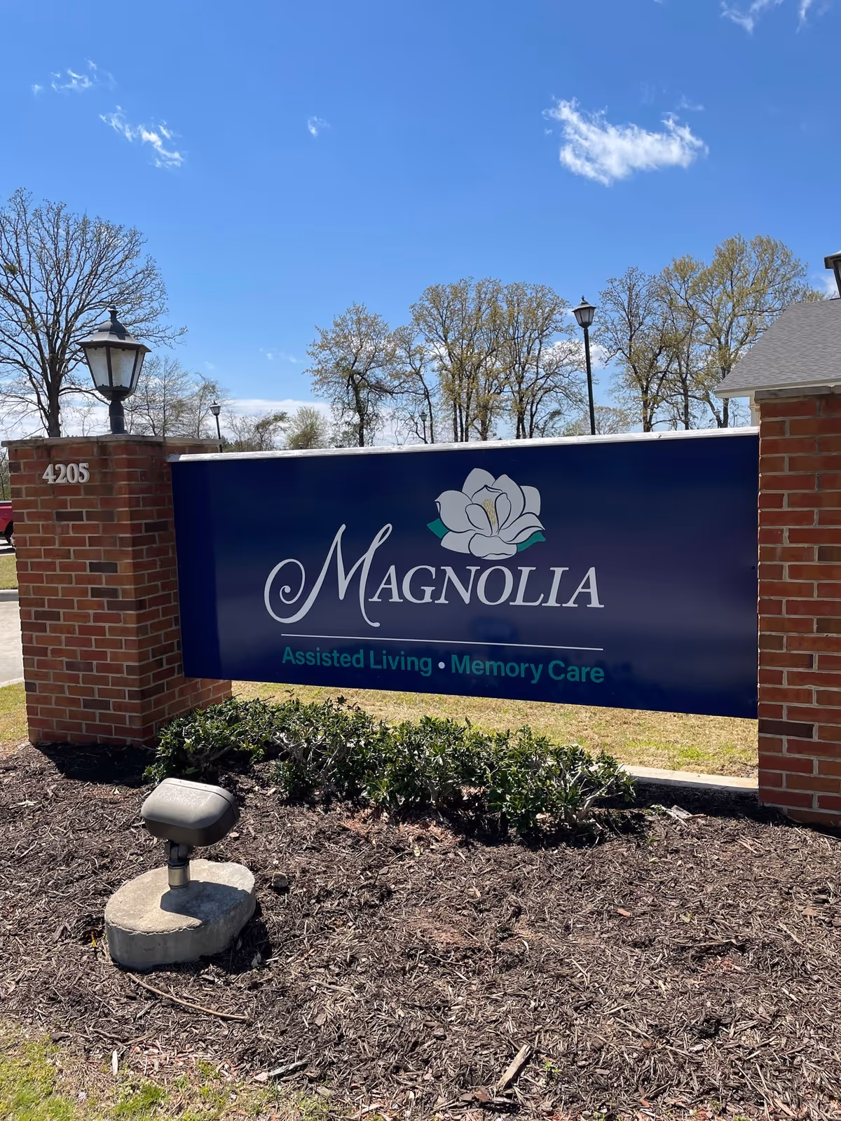 Outdoor view of a Magnolia Assisted Living and Memory Care sign mounted between two brick pillars with a lamp on top of each pillar. The sign is blue with white and teal text and a magnolia flower logo. Trees and a clear blue sky are visible in the background.