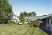 Exterior view of a single-story assisted living facility building with a green lawn, trees, and shrubs under a clear sky.