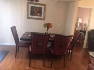 Dining area with a rectangular dark wood table set with placemats and a centerpiece of flowers. Four dark wood chairs with red cushions surround the table. A framed floral painting hangs on the wall above the table. Hardwood flooring and a doorway leading to another room are visible.