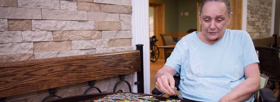 An elderly woman in a light blue shirt sitting in a wheelchair, working on a jigsaw puzzle on a table in front of her. The background shows a stone wall and a wooden bench, with a room and furniture visible further behind.