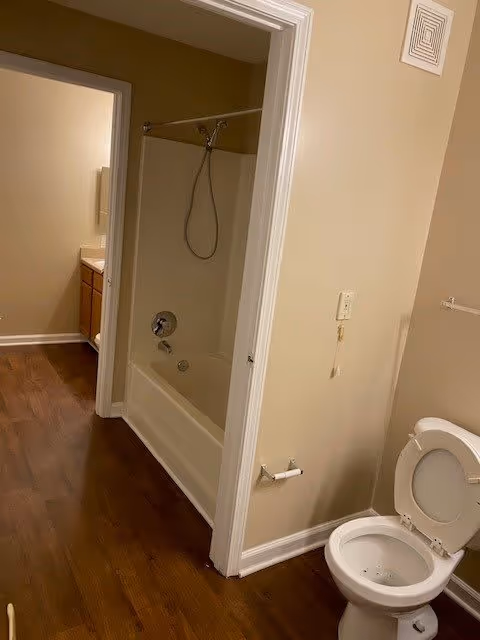View of a bathroom with a white toilet with the lid open on the right, a bathtub with a showerhead and curtain rod in the center, and a sink with a mirror visible through an open doorway on the left. The floor is wood, and the walls are beige.