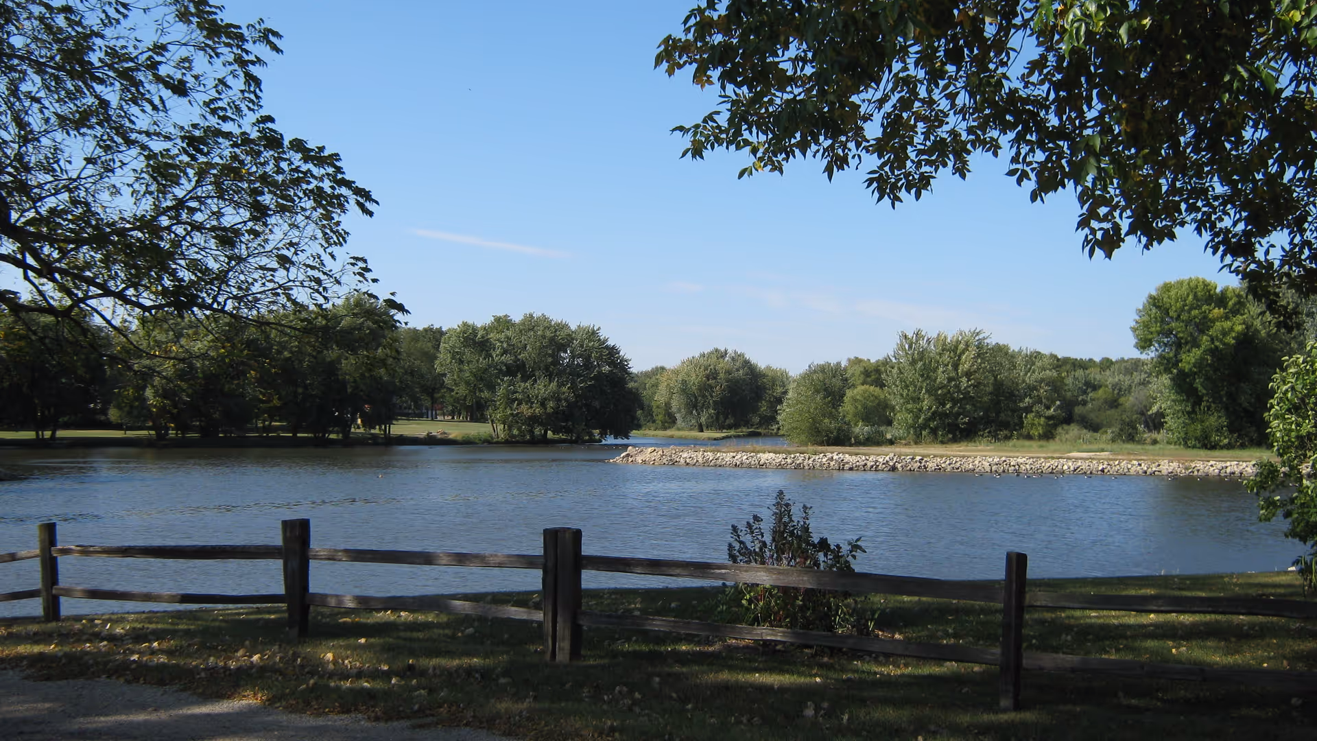 A peaceful outdoor scene featuring a calm body of water bordered by a wooden fence in the foreground and lush green trees in the background under a clear blue sky.