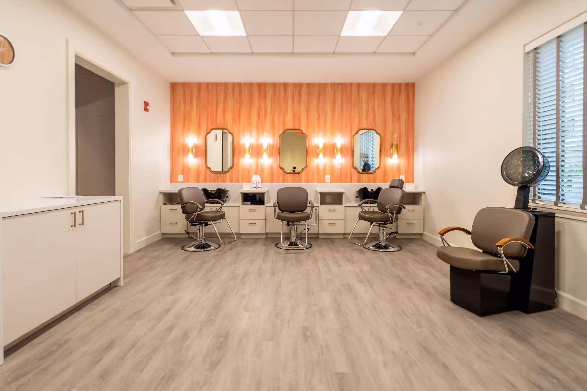 Interior view of a salon area with three styling chairs in front of a wall with four mirrors and wall-mounted lights. There is a hair dryer chair near a window with blinds on the right side and a cabinet on the left side. The floor has light wood-like flooring and the walls are painted white with an accent wall behind the mirrors.