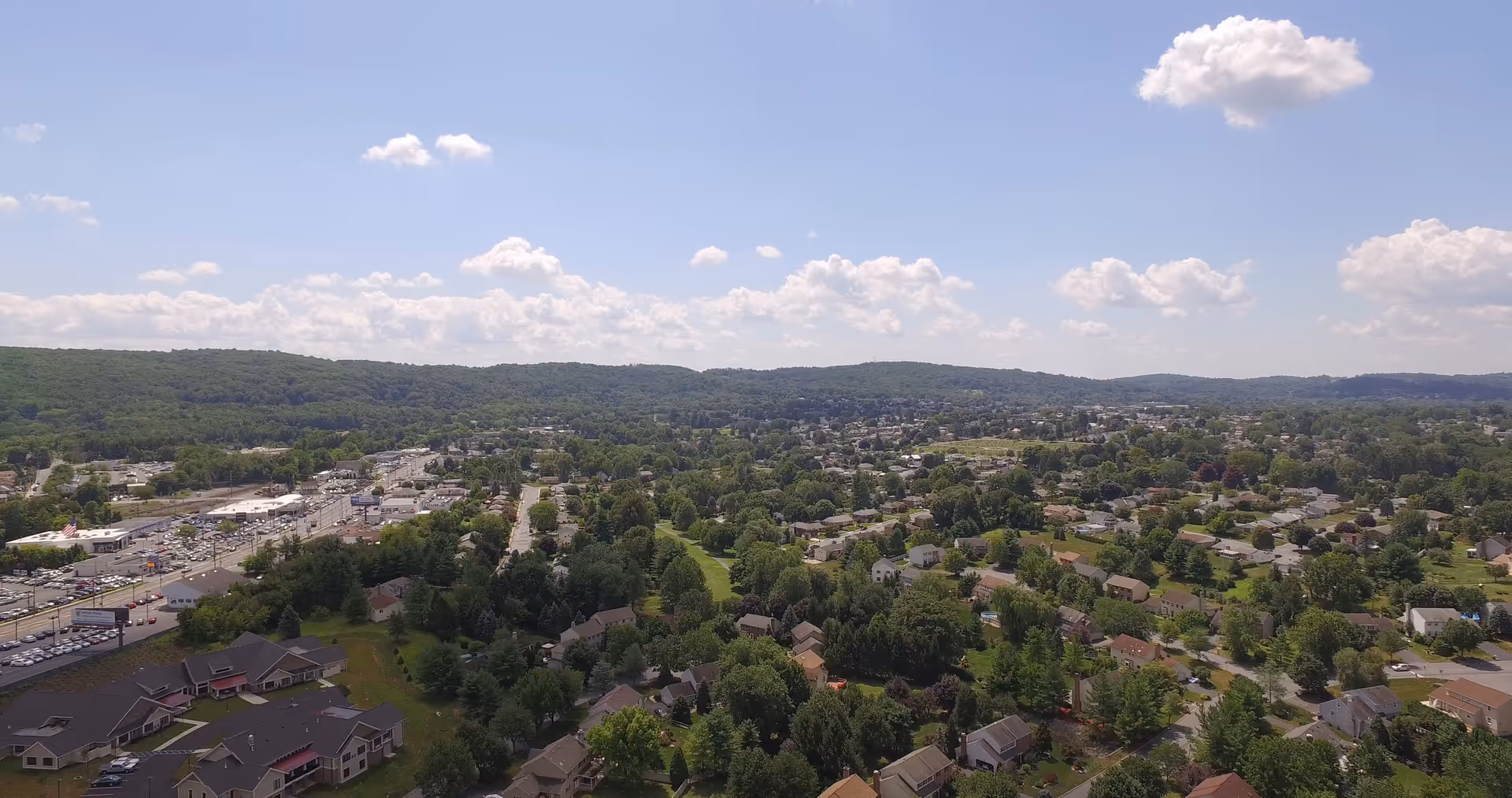 Aerial view of a suburban neighborhood with houses, trees, a commercial strip, and distant hills under a partly cloudy sky.
