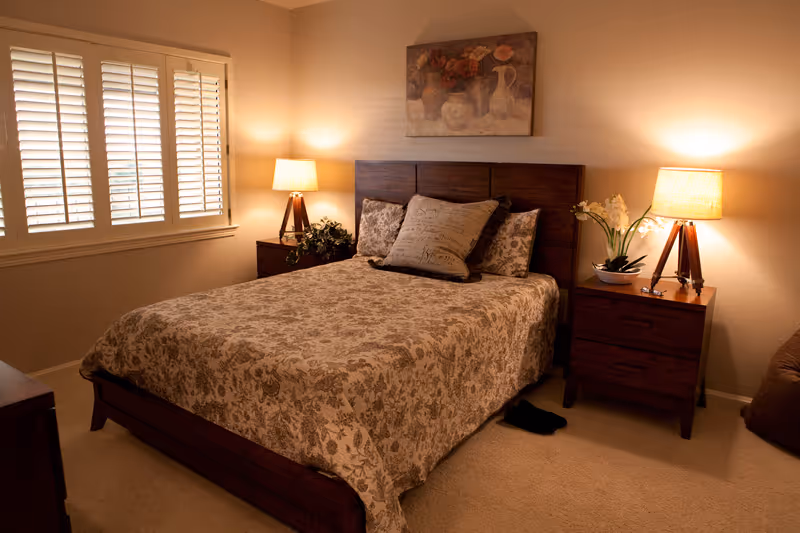 A warmly lit bedroom with a patterned bed, two wooden nightstands and lamps, a shuttered window, and artwork above the headboard.