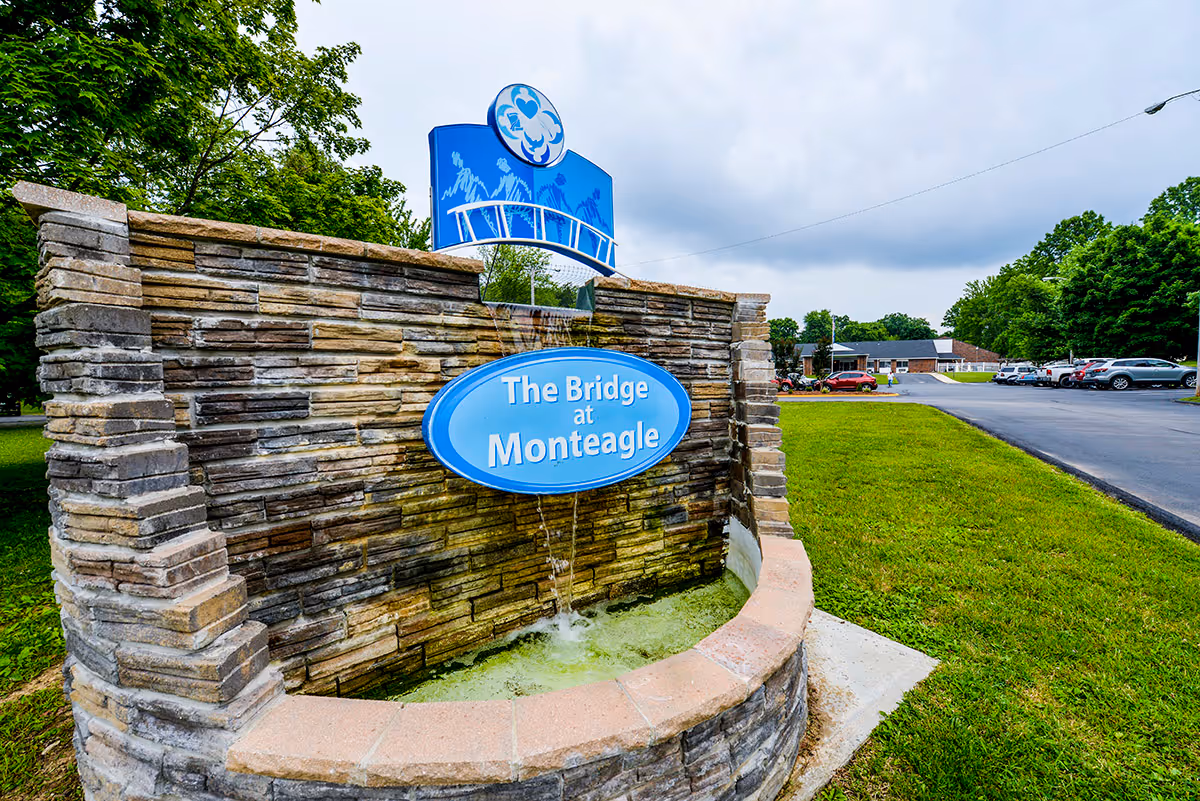 A stone fountain and blue sign reading "The Bridge at Monteagle" with a parking lot, building and trees in the background.