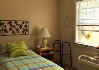 A small bedroom with a single bed covered in a striped bedspread with green, blue, and beige colors. There is a green pillow on the bed. Next to the bed is a wooden nightstand with a beige lamp, a small clock, and a few items. A black walker is positioned near the nightstand. A window with blinds lets in natural light, and there is an air conditioning unit below the window. The walls are beige, and a decorative metal wall art piece hangs above the bed.
