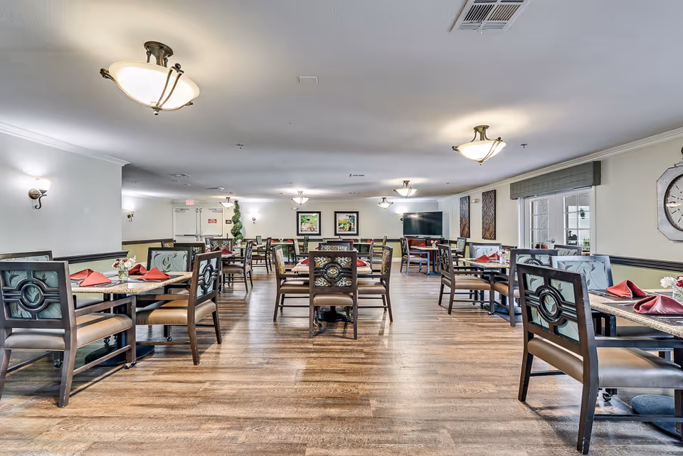 A spacious dining room with multiple tables and chairs arranged neatly. Each table has a placemat, a small flower vase, and folded red napkins. The room has wooden flooring, light-colored walls, ceiling lights, and wall sconces. There are framed pictures on the far wall and a large TV mounted on the right side. Windows with blinds are visible on the right wall.
