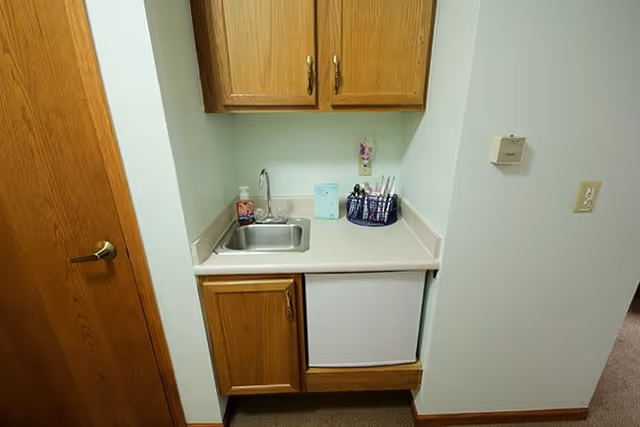 A small kitchenette area with a stainless steel sink, wooden cabinets above and below the countertop, a small white refrigerator under the counter, and a container holding utensils on the countertop. The walls are light-colored and there is a wooden door to the left.