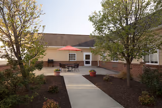 Outdoor patio area at Tyler's Retreat at Iron Bridge with a concrete walkway leading to a seating area featuring a table with a red umbrella and chairs, surrounded by trees and landscaping.