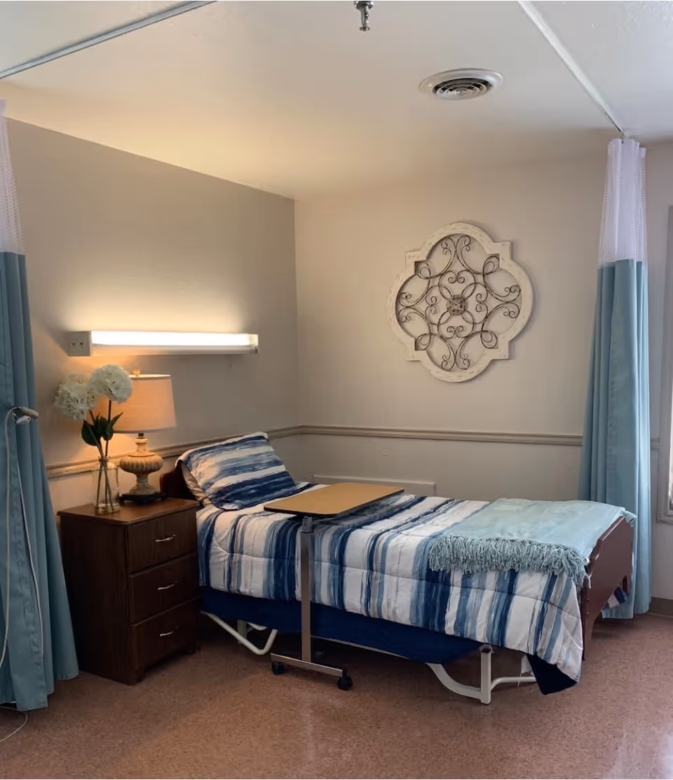 A neatly made single bed in a senior care room with blue-striped bedding, an overbed table, nightstand with a lamp and flowers, and decorative wall art.