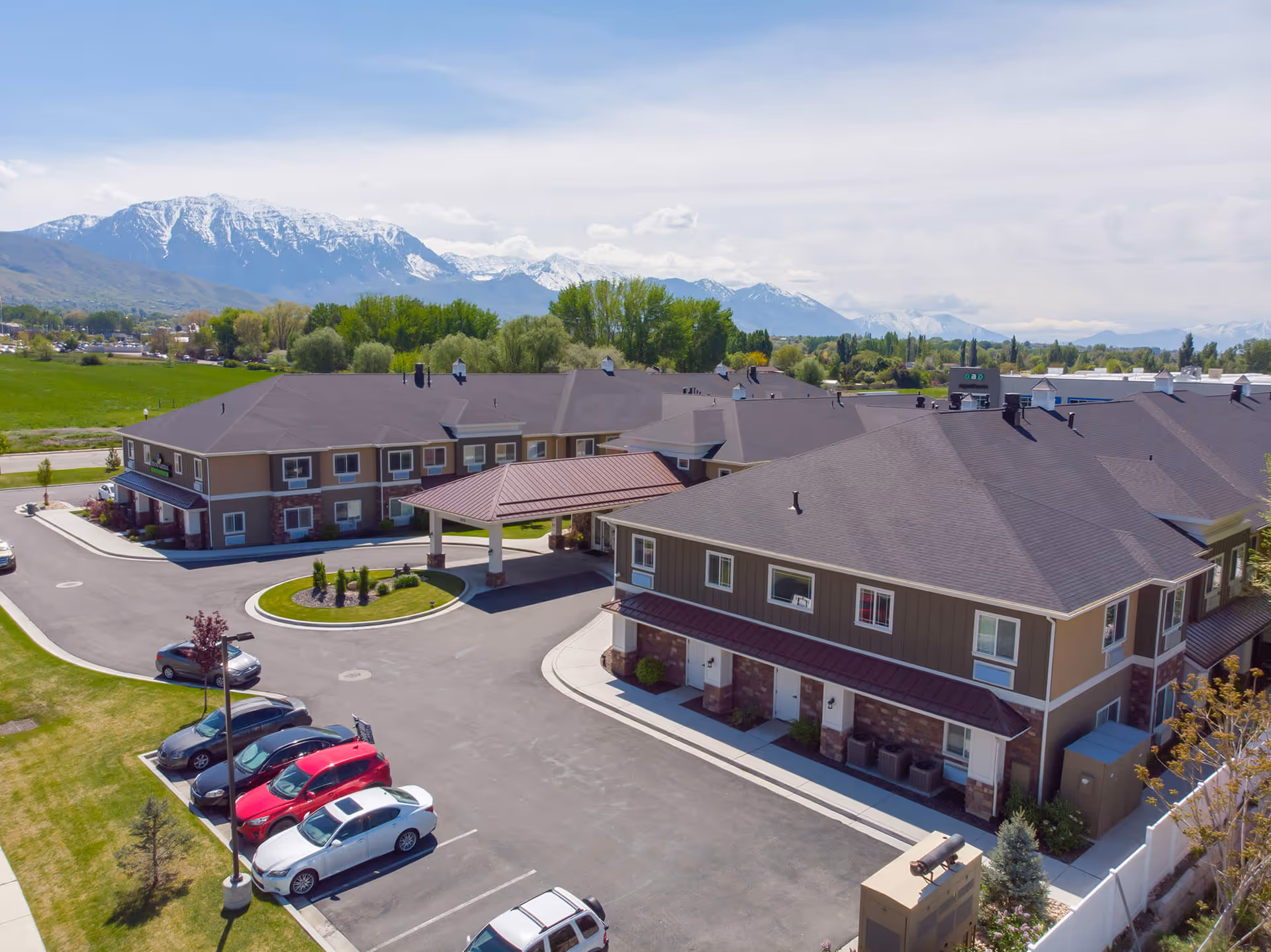 Aerial view of Spring Gardens Senior Living Lindon facility showing multiple two-story buildings with brown roofs and beige walls, a covered entrance, a parking lot with several cars, green lawns, and trees. Snow-capped mountains and a partly cloudy sky are visible in the background.