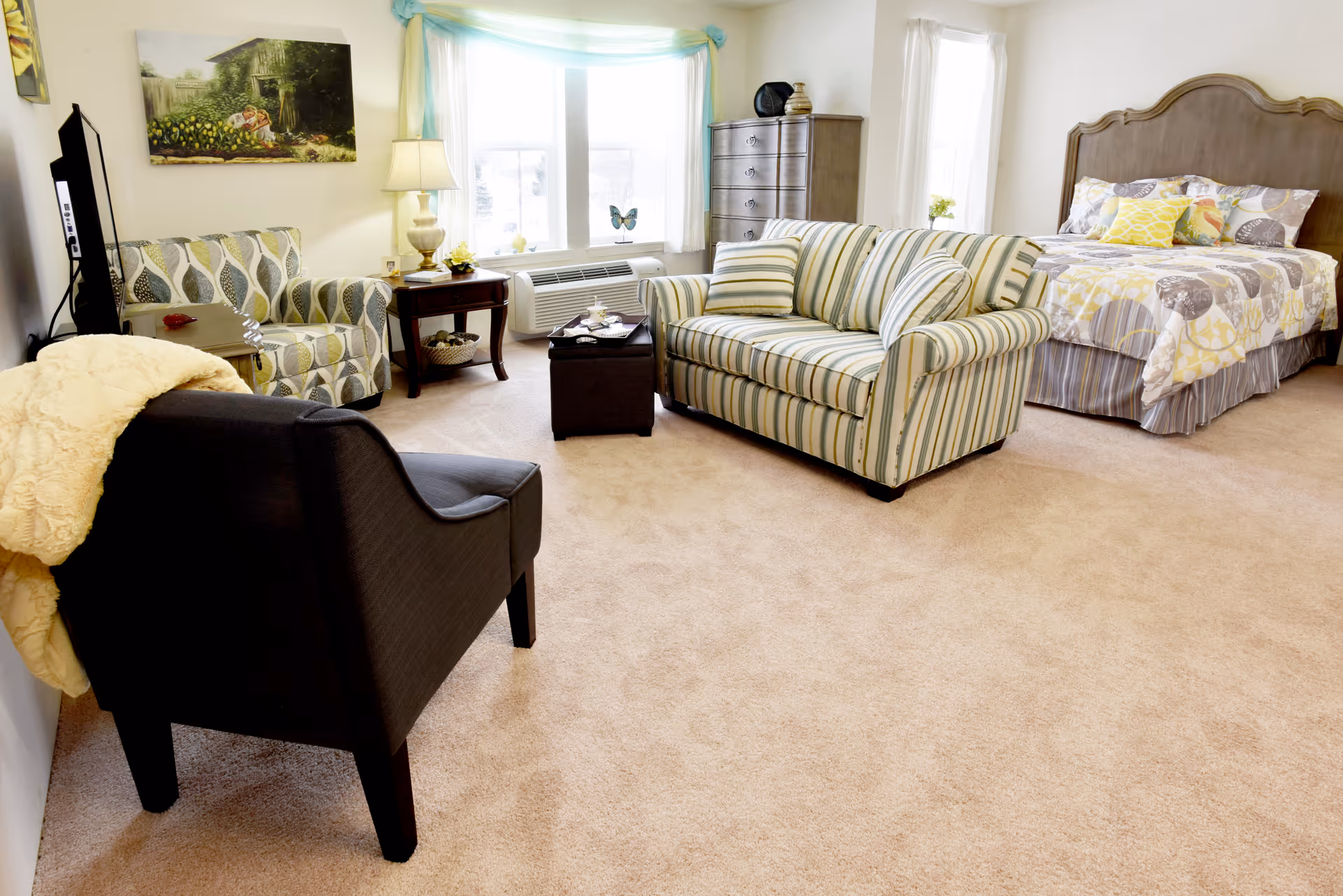 A bright and cozy room featuring a sitting area with a striped sofa, patterned armchair, and a dark armchair with a light-colored throw. There is a wooden side table with a lamp and decorative items, a large window with sheer curtains, and a bed with a wooden headboard and patterned bedding in soft yellow and gray tones. A tall dresser stands near the window, and the room is carpeted in a light beige color.