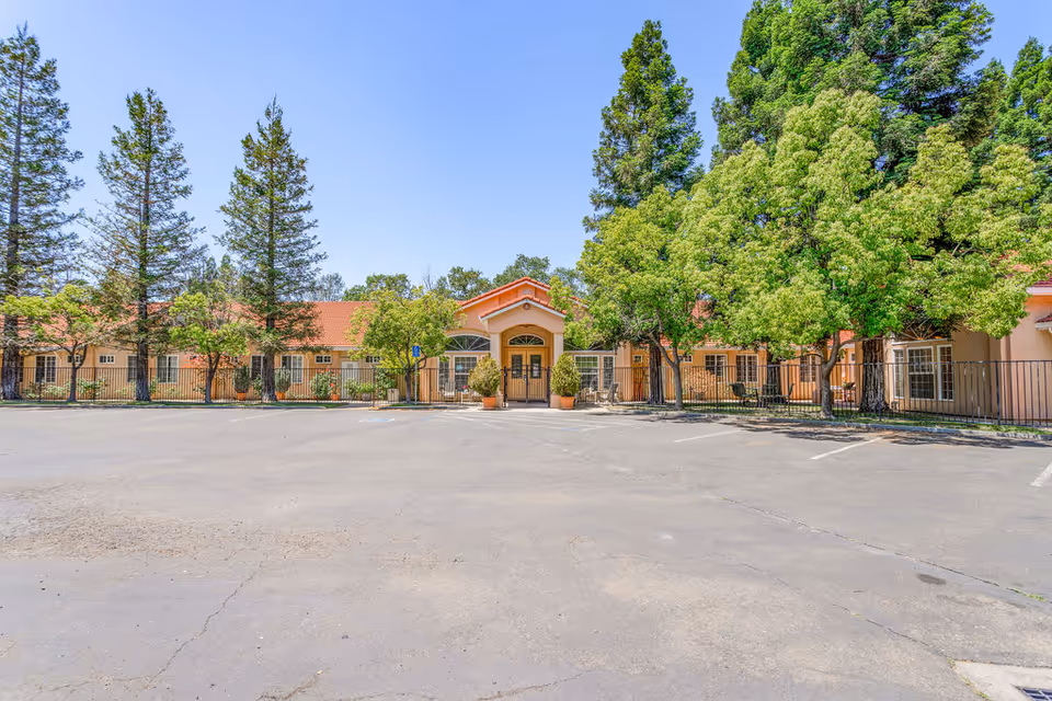 Front exterior view of a senior living facility with a wide driveway and parking area in front. The building has a light peach-colored facade with a red-tiled roof, surrounded by tall green trees and some smaller plants. The entrance is centered with a small covered porch and double doors.