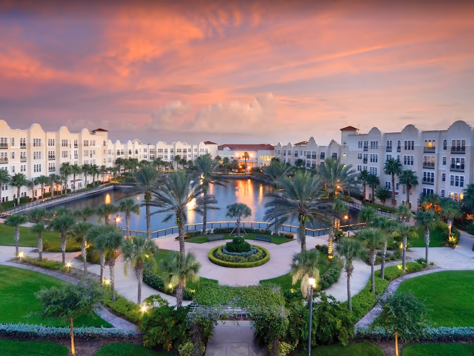 Aerial view of The Glenridge on Palmer Ranch senior living facility at sunset, featuring white multi-story buildings surrounding a central pond with palm trees and landscaped gardens, under a vibrant pink and orange sky.