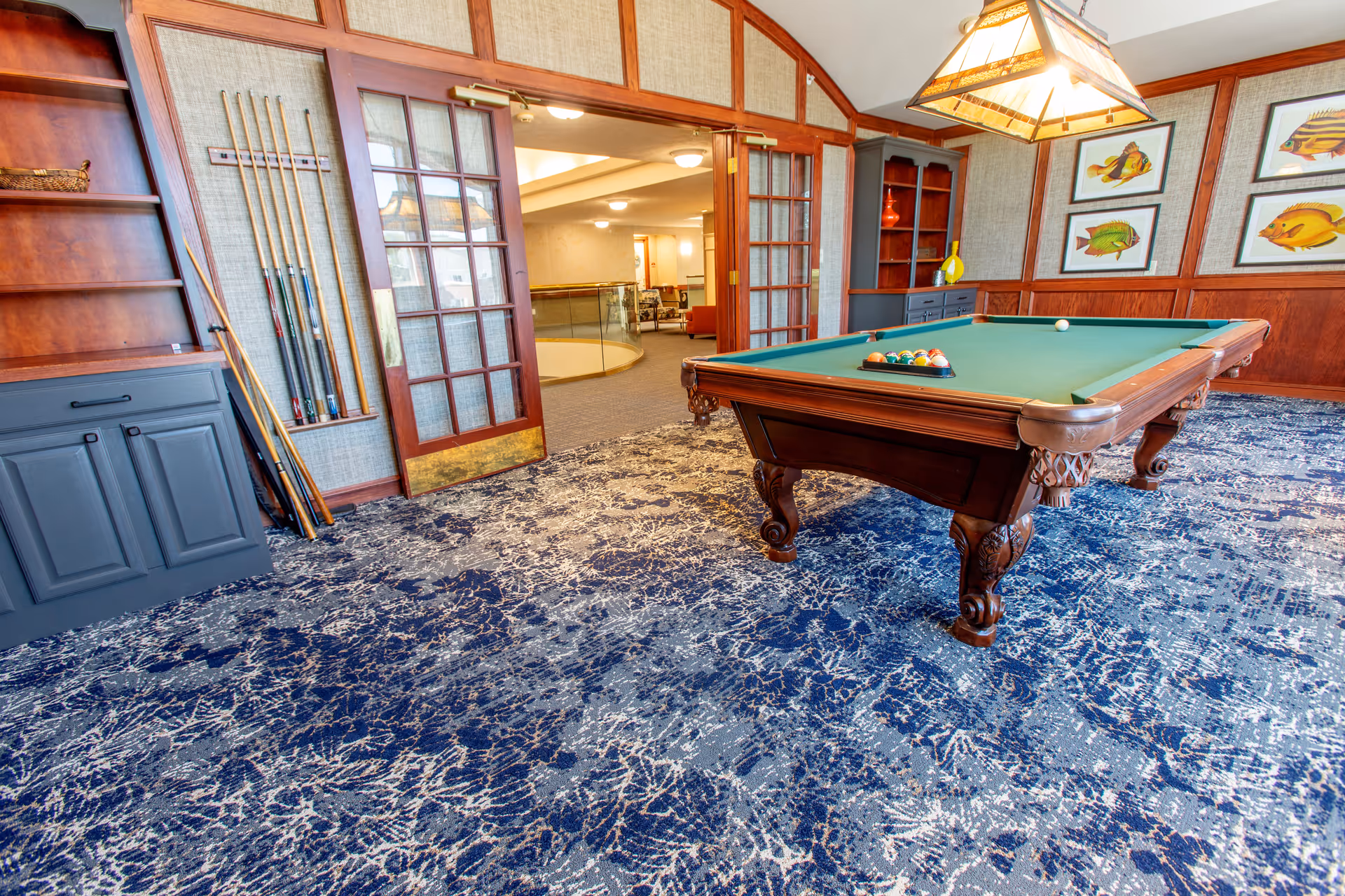 A recreational room with a pool table set up for a game, featuring a rack of pool cues on the wall and a wooden cabinet. The room has patterned blue carpet, wood-paneled walls with framed fish artwork, and a large hanging light fixture above the pool table. Double glass doors open to a hallway with seating visible in the background.