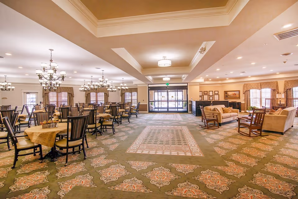 Spacious dining room with multiple round tables covered with beige tablecloths and surrounded by wooden chairs. The room features patterned carpet, chandeliers hanging from the ceiling, large windows with curtains, and a seating area with sofas and armchairs near the entrance.