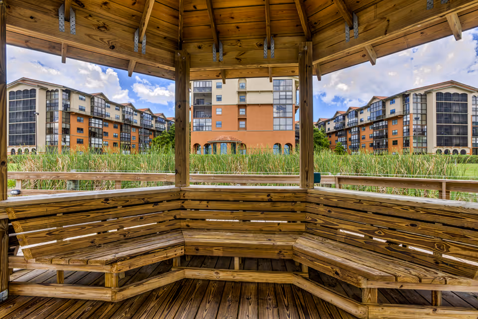 View from inside a wooden gazebo with built-in bench seating overlooking tall grasses and a multi-story residential building under a partly cloudy sky.