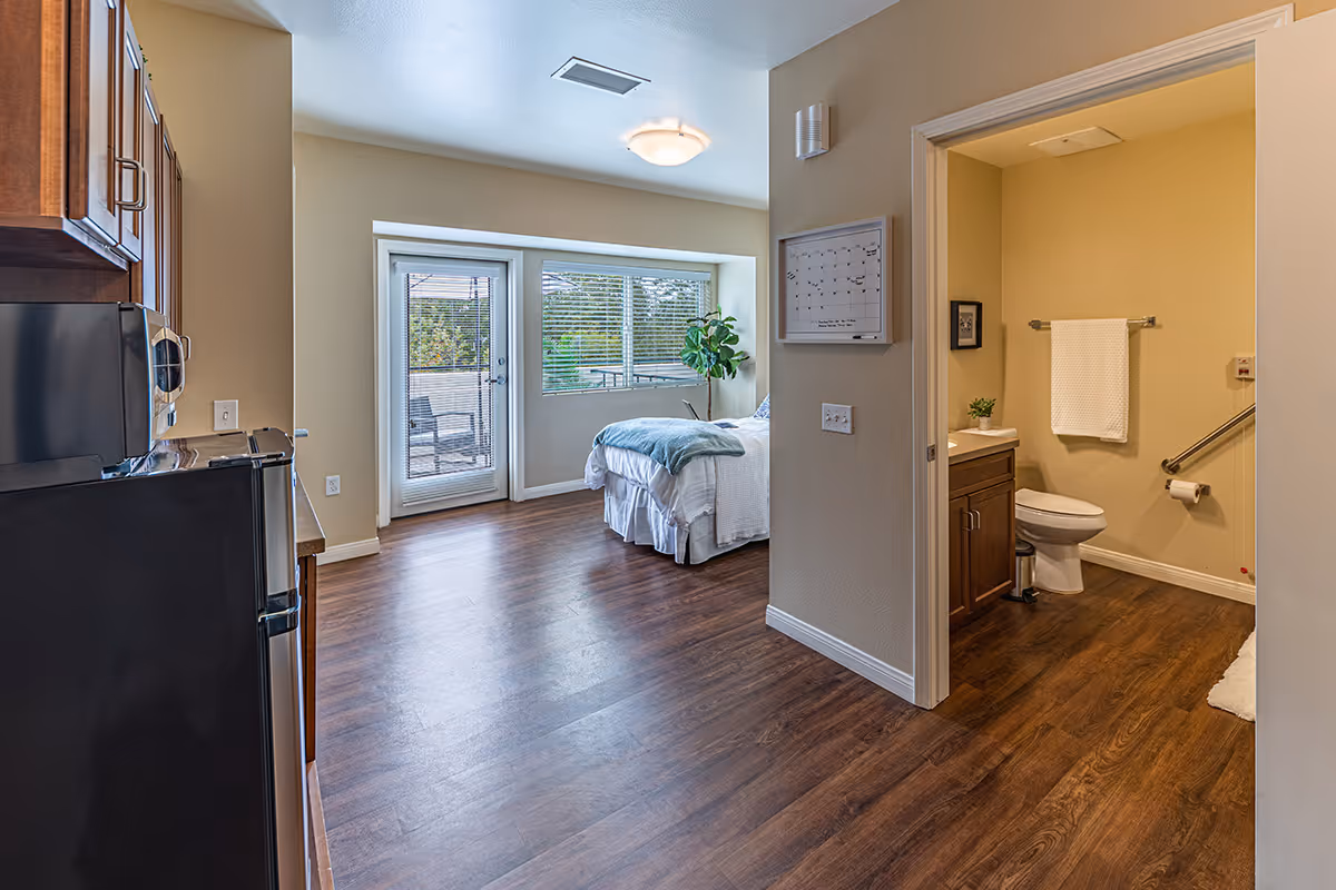Interior view of a senior living facility room showing a small kitchen area with a refrigerator and microwave on the left, a bedroom with a bed and a large window with blinds in the center, and a bathroom with a toilet, sink, and towel rack on the right. The room has wood flooring and beige walls, and there is a door leading to an outdoor patio visible through the window.