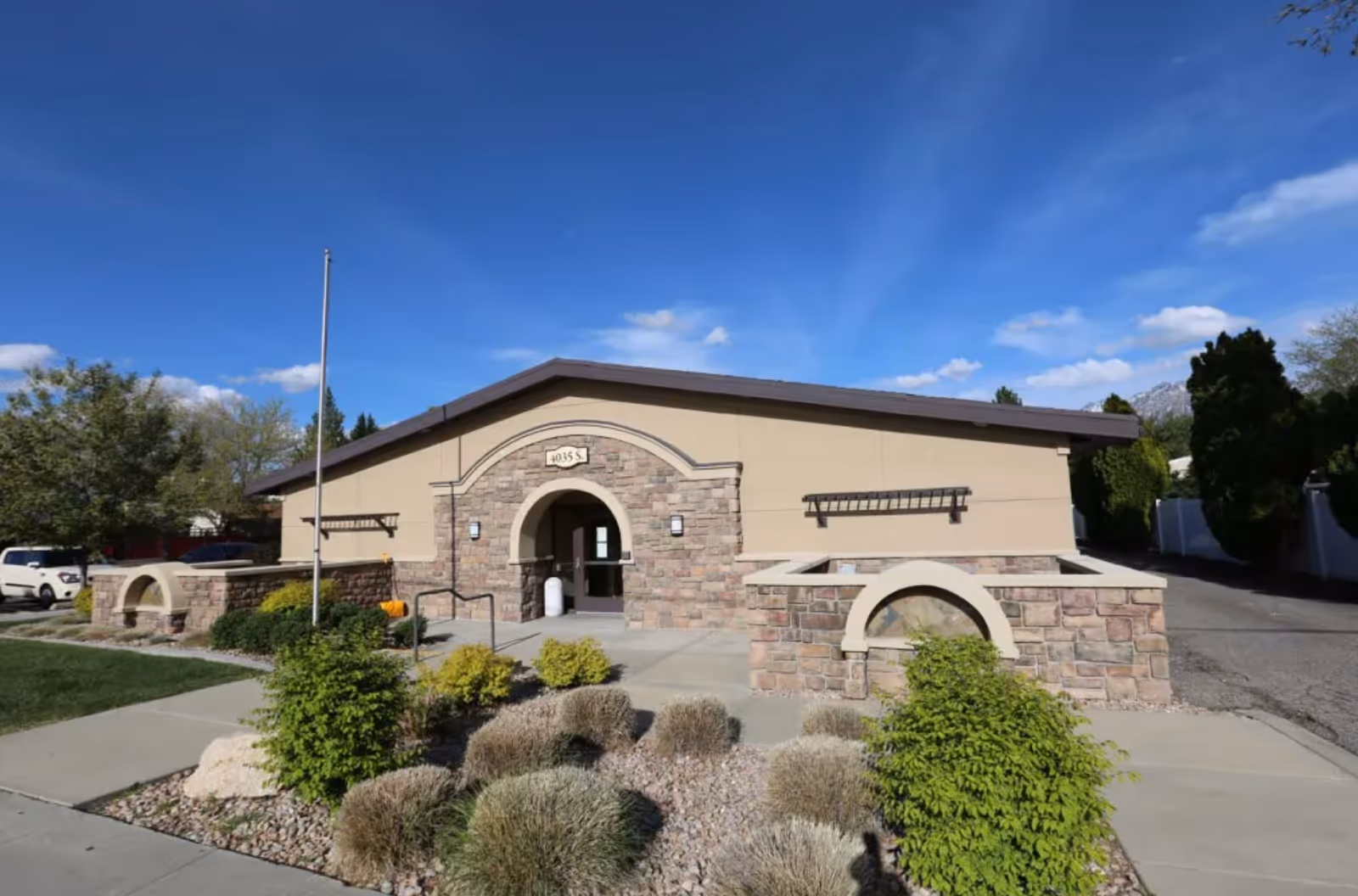 Exterior view of a single-story building with a stone and beige facade, an arched entrance, and a landscaped front area with bushes and rocks under a clear blue sky.