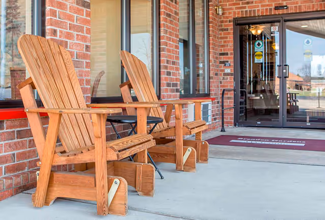 Two wooden Adirondack chairs and a small black metal table on a concrete patio outside a brick building with glass double doors and windows.
