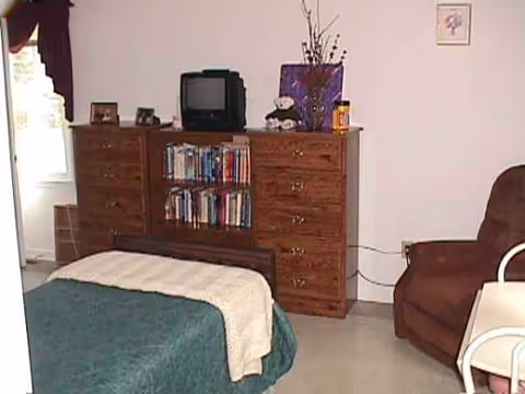 A bedroom in a senior living facility with a bed, dresser, TV, and chair.