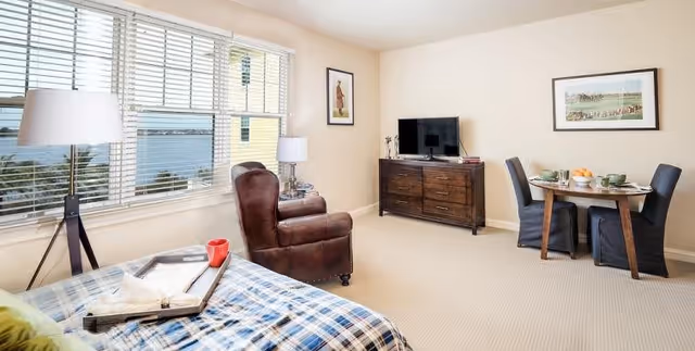 A bright and cozy room featuring a bed with a blue plaid blanket, a brown leather armchair, a wooden dresser with a flat-screen TV on top, a small round dining table set for two with green dishes, and large windows with white blinds allowing natural light to fill the space.