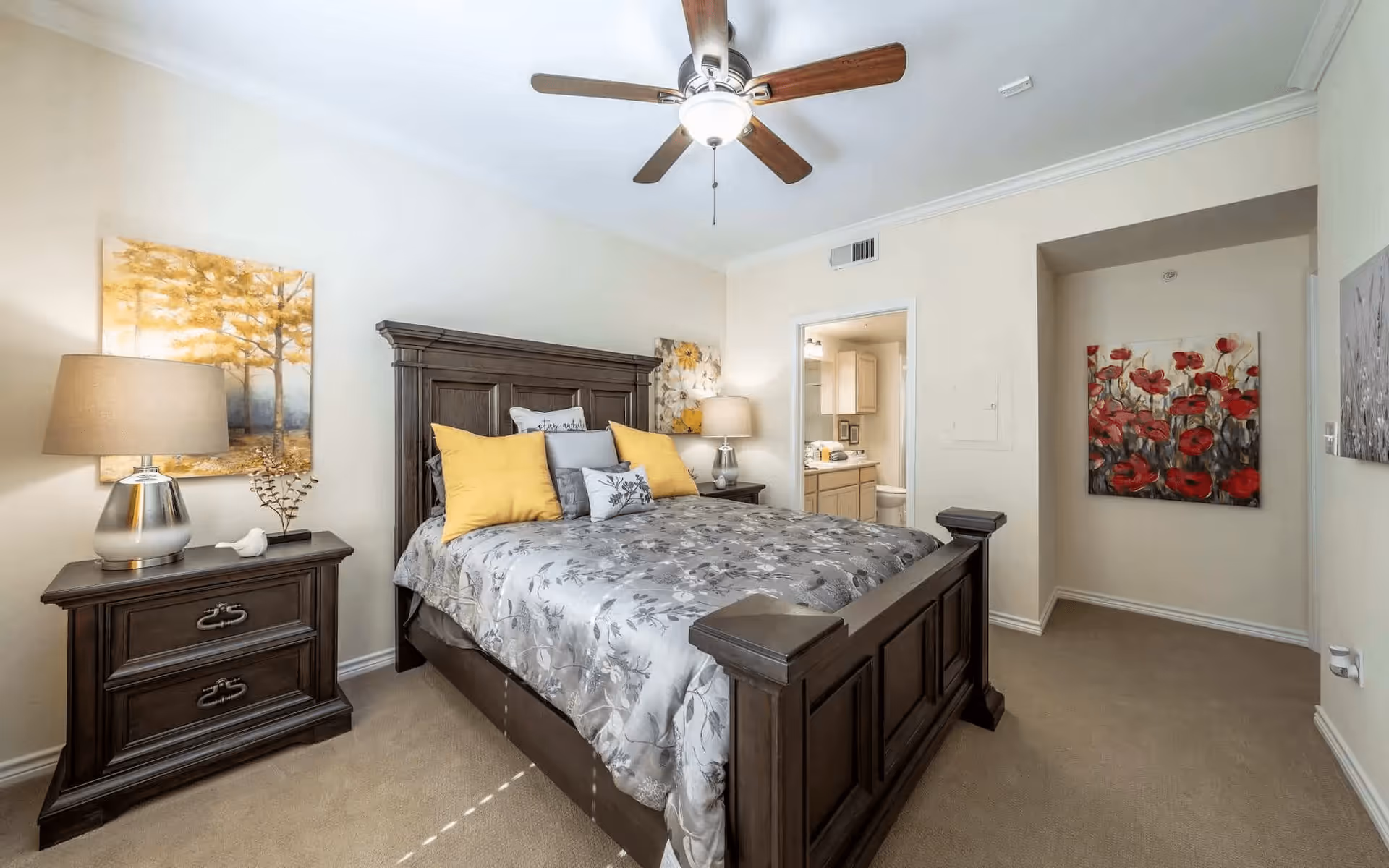 A well-lit bedroom with a large dark wooden bed frame and a floral patterned bedspread. The bed has several pillows, including two yellow ones. There are two matching dark wooden nightstands on either side of the bed, each with a lamp. The room has light-colored walls adorned with paintings, one depicting yellow trees and another with red flowers. A ceiling fan with wooden blades is mounted on the ceiling. An open doorway leads to a bathroom with light cabinetry and a toilet visible.