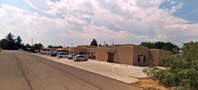 Exterior view of a single-story building with a flat roof and beige stucco walls, a parking lot with several parked cars, and some trees and shrubs around the property under a partly cloudy sky.